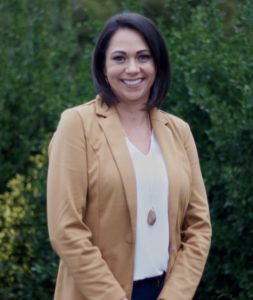 A woman in a tan jacket standing next to bushes.